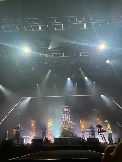The image shows a stage at the International BCA Java Jazz Festival in Salembaran, Indonesia. A band is performing, featuring musicians on keyboards, drums, guitar, and other instruments. The stage setup includes a lighting grid above and rows of lights behind the performers. The stage is illuminated with spotlights.