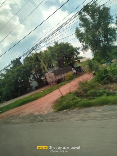 The image shows a slightly tilted view of a street scene in Ben Cau, Vietnam. A dirt road runs diagonally through the frame, bordered by green grass and lush trees on either side. A large truck with a covered bed is parked on the dirt road, with a white structure behind it. Power lines are strung across the sky, intersecting with the branches of the trees. The sky is partly cloudy, with a mix of blue sky and white clouds. The overall impression is of a rural or suburban area on a cloudy day. The timestamp in the lower right corner indicates the image was taken on June 24, 2026, at 09:48.