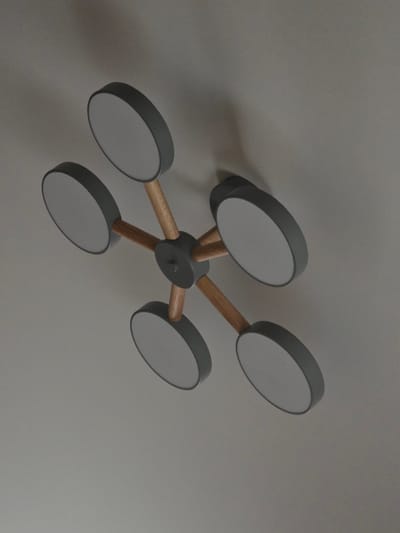 The image shows a close-up, high-angle view of a modern ceiling light fixture. The fixture has a central, dark gray circular hub from which five wooden arms extend outwards. Each arm terminates in a circular, gray-rimmed light panel with a white diffuser. The light fixture is mounted on a plain, light gray ceiling. The image does not contain any information about the city of Ulyanovka, Russia, nor does it depict any people or actions. It is a static, interior shot focusing solely on the design of the lighting fixture.