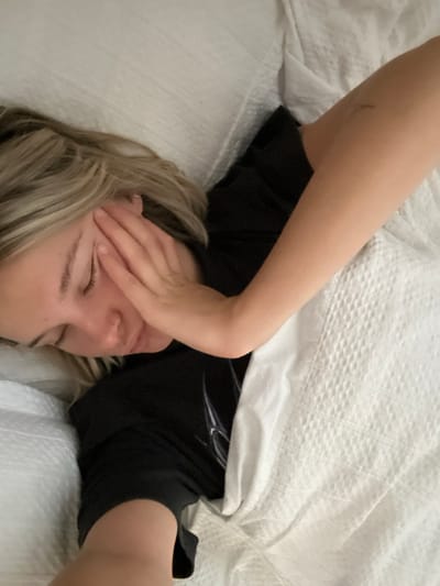 The image displays a Caucasian female lying on her right side in a bed, appearing to be asleep or resting. Her head rests on a white pillow, with her blonde hair spread on the pillow and her eyes closed. A small silver nose ring is visible in her left nostril. She wears a black short-sleeved shirt featuring a faint, light-colored graphic design on the left chest area. Her right hand is positioned over the left side of her face, covering her left eye and temple. Her body is partially covered by a white, textured duvet or blanket. A small, faint mark is present on her right upper arm. The setting is an indoor private space, consistent with a bedroom. No visual information within the image definitively identifies the location as Victoria, Canada.
