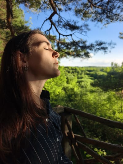 A young woman is captured in a profile view, facing right with her eyes closed and head tilted slightly upwards, basking in the sun. She has long, dark hair and wears a dark blue striped shirt with white buttons, a silver hoop earring in her left ear. The background reveals a lush, green landscape of trees and foliage under a bright blue sky with some tree branches in the foreground. A wooden railing is visible in the lower right section of the image. The image appears to have been taken outdoors, possibly in a park or natural area, in the vicinity of Onokhino, Russia, given the description of the location.