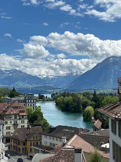 This is an eye-level shot overlooking a body of water, likely a lake or river, in Thun, Switzerland. The water is a vibrant turquoise color and is bordered by lush green trees and vegetation. In the foreground, there are several buildings with red-tiled roofs, characteristic of European architecture. One building on the left has a striking half-timbered facade. Further back, a grand, multi-story building with a dark roof and many windows stands prominently near the water's edge. Beyond this, a wide expanse of water leads towards a range of majestic mountains with snow-capped peaks under a bright blue sky filled with fluffy white clouds. A small Swiss flag is visible on top of the grand building. The scene appears to be a scenic view of the Alps from Thun, with the city's rooftops and a prominent building framing the natural landscape.