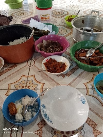 The image shows a table set for a meal in an Indonesian home in Gegesik, located at coordinates -6.538 latitude and 108.43 longitude.  The table is covered with a brown and cream tiled surface. There are various dishes on the table, including a pot of white rice, a bowl of fried fish, a bowl of red chili paste, a bowl of fried tempeh, and a bowl of white rice with tempeh.  There is also a large empty plate and a smaller bowl that contains white rice with tempeh. There is a spoon on the table next to the smaller bowl.  In the upper left corner of the image is a small bowl with some leafy green vegetables. In the upper right corner of the image are two bowls, one containing a red sauce and the other containing a darker sauce. There is also a pot of water in the background. The scene suggests a meal is about to be served.
