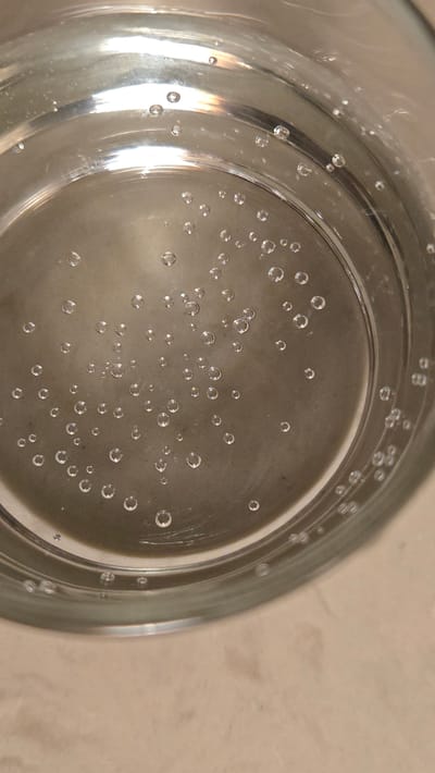 This media file is a close-up, top-down shot of a clear glass filled with a bubbly liquid, likely carbonated water or soda. The glass has a thick rim and appears to be made of glass. Numerous small, clear bubbles are suspended throughout the liquid and adhere to the inner surface of the glass. Some bubbles are larger and more defined, while others are tiny specks. The liquid itself is clear and reflects light, creating bright highlights on the glass's surface, particularly on the left side, suggesting a light source. The background appears to be a neutral, textured surface, possibly a countertop or table. There is no visible text in the image. The scene is static, focusing solely on the visual characteristics of the carbonated beverage within the glass. The overall impression is one of refreshment and clarity, with the bubbles adding a sense of effervescence. The lighting suggests an indoor setting, possibly during the day, though no specific time cues are discernible. The location context provided is Santa Ynez, United States of America, but this cannot be visually confirmed from the image itself.