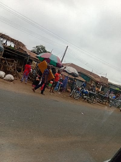 The image displays a street scene in Itobe, Nigeria, under an overcast sky. A paved asphalt road occupies the foreground and mid-ground. To the right, a dirt shoulder abuts a row of market stalls and utilitarian buildings. Approximately 10 to 15 individuals are visible within the scene. In the mid-foreground, a male subject in a striped shirt and dark trousers walks from left to right, carrying a large, rectangular yellow object, possibly a foam mattress or board, on his left shoulder.
Along the right side of the road, several market stalls are established. Two prominent market umbrellas are deployed, one multi-colored (red, green, light blue) and another white/grey, providing shade. White sacks, possibly containing produce, are visible on the ground near the leftmost stalls. Other individuals are congregated around these stalls; one person in a red top and patterned trousers is partially visible near the left side of the market area, while others are less distinct. At least four motorcycles are parked on the dirt shoulder in front of the market stalls. The buildings behind the stalls are generally single-story structures with corrugated metal roofs. Overhead utility lines and a wooden utility pole are visible against the light grey sky.