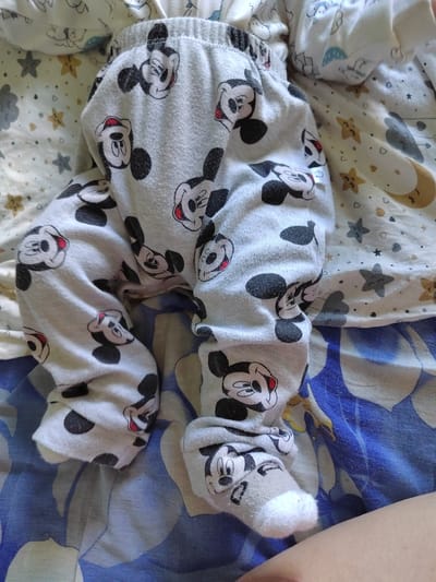 The image shows a close-up view of a baby's legs and feet, dressed in pajamas featuring a pattern of Mickey Mouse faces. The baby is lying down on a patterned surface, likely a bed or blanket. The pajamas are light gray with black and red Mickey Mouse prints. The baby's feet are covered with socks that also have a Mickey Mouse design. The background is a collection of patterned fabrics, one with stars and moons, and another with a blue and white swirling design. The scene appears to be indoors, possibly in a bedroom, given the presence of bedding. The lighting suggests daytime. There is no visible text in the image. The overall impression is one of comfort and a focus on the baby's attire.