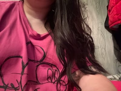 A close-up shot shows a person with long, dark hair wearing a pink shirt with a black design. The person's neck and shoulder are visible, along with a glimpse of patterned wallpaper and a red quilted item in the background. The location is not discernible from the image. There are no actions or interactions visible.