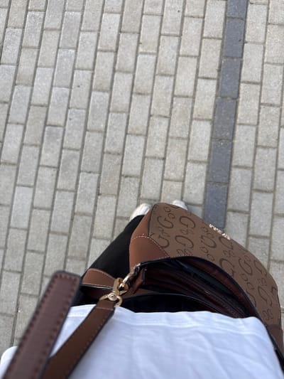 The image shows a first-person perspective of someone standing on a brick-paved sidewalk in Szklarska Poręba, Poland. The sidewalk is composed of light gray rectangular bricks with a darker gray strip running horizontally across it.
The subject's attire includes black pants and white sneakers. They are holding a brown handbag with a repeating "G&G" logo pattern in a lighter brown color. The handbag also features brown leather trim and a gold metal clasp. A white garment, possibly a shirt or jacket, is visible on the right side of the frame.
The scene appears to be outdoors during daylight, with natural lighting. There are no specific indicators of the time of day or weather conditions, other than the brightness suggesting it is not nighttime. The background is blurred, but the paving stones suggest an urban or semi-urban environment, possibly a street or a public walkway. The overall mood is neutral, focusing on the details of the clothing and accessories. There is no visible text other than the logo on the handbag.