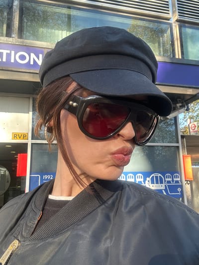A person is depicted in a close-up, slightly off-center profile shot under bright daylight. The individual wears a black newsboy-style cap, large black sunglasses with reflective reddish lenses and two silver studs on the side frame, a dark blue/black bomber jacket with a ribbed collar, and a layered grey and black top. A patterned hoop earring is visible on the left ear. The person's lips are pursed, forming a pucker. The background reveals the exterior of a London public transport station. Visible elements include a blue sign with white text "STATION" on the upper left, a red sign labeled "RVP", and a partial blue sign displaying "1992". A larger blue panel on the right features white graphics representing various London transport modes, specifically a DLR train, a London Underground roundel, and a bus icon. The station architecture consists of glass panels and metal framing. This scene is situated at the entrance of a London Underground or DLR station within the City of Westminster, United Kingdom.