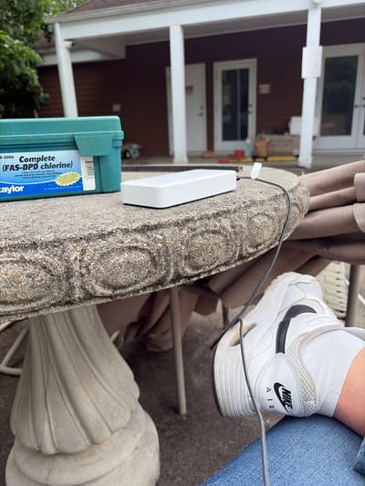 The image shows a close-up view, likely taken outdoors, with a combination of natural and man-made elements. A weathered, ornate concrete table is in the foreground, upon which a white electronic device and a blue container labeled "Taylor Complete (FAS-DPD chlorine)" are placed. A black cable is connected to the electronic device. A person wearing light blue jeans and white Nike Air sneakers with black swooshes is visible. The background shows a two-story house with white pillars and a brown exterior. There are green trees. The location is likely in Edison, United States of America.