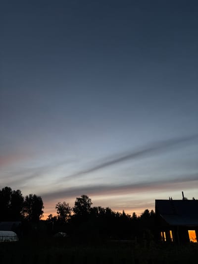 This is a vertical, nighttime photograph taken outdoors in Dubrovino, Russia. The scene captures a twilight sky with streaks of light, likely from clouds catching the last rays of the sun. On the right side of the image, a dark silhouette of trees borders the sky. A dark-colored building with illuminated windows is visible on the far right, suggesting indoor activity. The overall mood is serene and peaceful, indicative of the end of the day. The lighting suggests it is either dawn or dusk. No people are visible in the image, and there is no visible text.