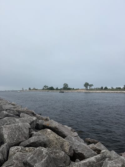The image shows a wide body of dark water with a rocky breakwater in the foreground. The water extends towards a distant, tree-lined shore with several boats visible on the water. The sky is overcast and pale gray. The scene is likely a harbor or channel entrance, and given the prompt, is situated in Muskegon, Michigan, United States of America. There are no people clearly visible in the image. The overall impression is one of a calm, overcast day at a waterfront location.