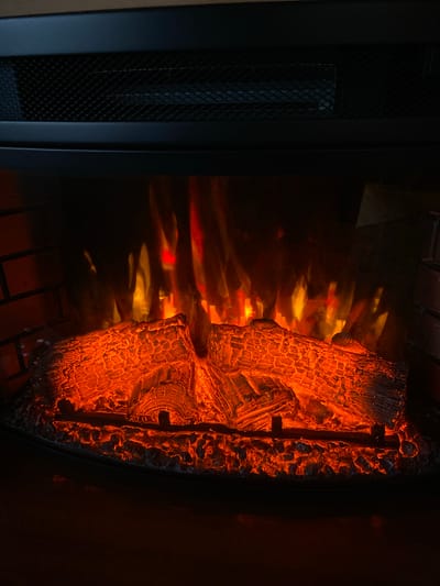 The image shows a close-up view of an electric fireplace with faux logs and glowing embers, set against a backdrop of simulated flames. The fireplace has a black metal grate at the top, and a brick-like texture on the left side of the frame. The logs are detailed, showing the texture of bark and charred wood, and they are illuminated by a warm, orange and red glow from the embers. The flames appear to be a mixture of reds, oranges, and yellows, creating a sense of warmth and ambiance. The surrounding environment appears to be dark, likely indicating that the room is dimly lit or the fireplace is the primary light source. There are no people or animals present in the image. The scene depicts a man-made simulation of a fire, designed for decorative purposes. The location in the city of Vysokiniči, Russia, is not discernible from the image itself.