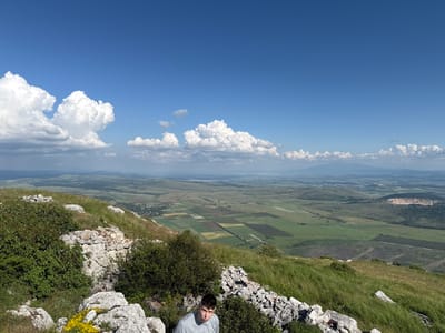 The image presents a panoramic view from a high vantage point, overlooking a vast landscape under a clear blue sky with scattered white cumulus clouds. In the foreground, a steep, rocky hillside covered in green grass and sparse bushes occupies the lower portion. Visible among the rocks on the left are small clusters of yellow wildflowers. A young male with short dark hair, wearing a light grey t-shirt, is positioned in the lower-center, partially framed by rocks and vegetation, looking directly towards the viewer. The midground unfolds into an expansive plain, characterized by a distinct patchwork of agricultural fields in various shades of green and brown. A small village with clustered buildings is discernible on the left side of the plain, while a large open-pit quarry with exposed light-colored earth is prominent on the right. In the distant background, hazy mountains or hills stretch across the horizon. This scene is situated in the area of Dragoman, Bulgaria.