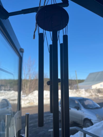 The scene, located in Сундур, Russia, is observed from an interior looking outward through a glass panel. A dark-colored wind chime is prominently displayed in the foreground, suspended by twine from a dark metal frame, likely a door or window frame. The wind chime features a circular wooden top piece from which multiple dark, cylindrical tubes of varying lengths hang via black cords. A horizontal striker is positioned within the tubes. The exterior environment is illuminated by a clear, bright blue sky. Snow covers much of the ground, forming drifts and patches, interspersed with darker areas of exposed earth or pavement. A light-colored, compact hatchback car, possibly silver or light gray, is partially visible on the right side, positioned on or near a road. In the mid-distance, bare deciduous trees and a distant line of evergreen foliage are present. To the right, a light-colored building with a gray, sloped roof and a utility pole are also visible in the background.