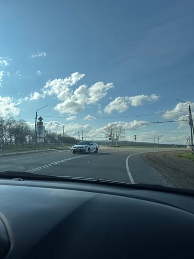 A photo taken from inside a car shows a view of a road with a silver car driving on it. On the left side of the road, there are trees and power lines. The sky is blue with white clouds. The location appears to be Otradnaya, Russia based on the presence of roadside infrastructure and the overall landscape.