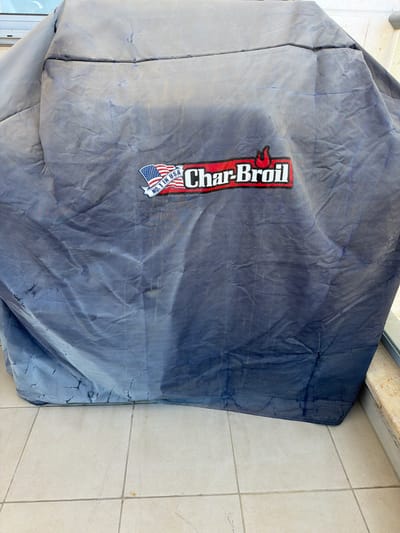 A dark blue grill cover with the "Char-Broil" logo is draped over an unseen object. The cover is wrinkled and appears to have water stains or discoloration. It is situated on a light-colored tiled surface, likely outdoors. The background suggests an urban environment, possibly a balcony or patio area, with a hint of a window frame and a beige stone ledge visible. The image was taken in Sderot, Israel.