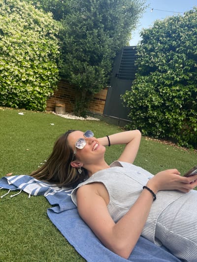 A young woman with long brown hair, wearing sunglasses, a sleeveless white and blue striped shirt, and a black bracelet, lies on a blue and white striped towel on the grass. She is looking up with a smile and is holding a black mobile phone. There are white flowers on the bushes bordering a brick wall to the left of the woman. The grass is green and the sky is visible through the foliage. The location of the scene is a backyard in Majadahonda, Spain, at the coordinates 40.469 latitude and -3.857 longitude. The woman's right arm is extended and she is holding a phone. Her left hand is resting on her head. She is wearing a thin gold chain necklace, and her left ear is pierced. The woman's legs are covered by the towel. There is a tall green bush to the right of the woman, and a gray slatted gate to her left. The ground is made of artificial grass.