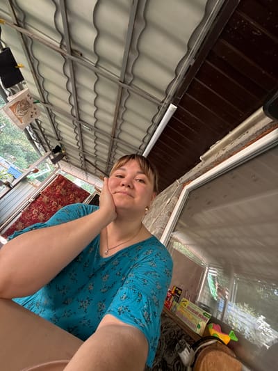 A selfie taken by a woman, likely outdoors in a rustic or backyard setting. The woman, with short brown hair and fair skin, is wearing a blue floral print dress and a delicate necklace. Her left hand is resting on her chin, and she is looking at the camera with a gentle, possibly contemplative expression.
The background reveals a corrugated metal roof overhead, suggesting a covered outdoor area such as a porch or patio. To the right, a window reflects the surrounding greenery and trees, indicating it is daytime and likely pleasant weather. Various objects are visible near the window, including what appear to be boxes of dominoes or board games, a toy, and some electrical cords. A reddish-brown patterned rug is draped in the background, adding a touch of domesticity to the scene. On a clothesline to the left, a white cloth with a tiger illustration and some dark clothing items are hanging, perhaps drying in the sun. The overall impression is one of a relaxed, informal moment captured in a semi-outdoor living space. The specific location of Nizhne-Chufichevo, Russia, provides further context for the type of environment depicted.