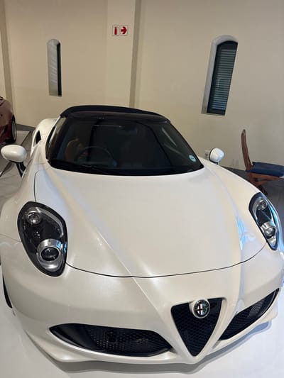 A white Alfa Romeo 4C sports car is parked indoors. The location appears to be a garage or showroom, judging by the smooth walls and window shape. The car is angled towards the viewer. The lighting is bright and overhead. There is no indication of the city of Stellenbosch Local Municipality or South Africa.