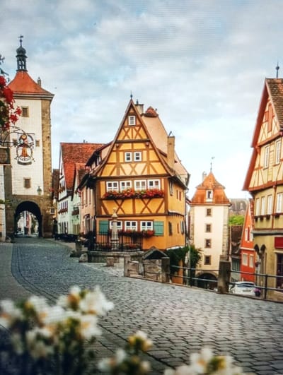 The image displays a street scene located in Legnica, Poland, characterized by historic, multi-story buildings and a cobblestone street under an overcast sky. In the immediate foreground, out-of-focus white and yellow flowering plants are visible. The dark grey cobblestone street, featuring a darker central strip, curves gently uphill towards the right.
To the left, a tall, light-colored tower structure with a dark orange conical roof, topped by a dark weather vane, stands adjacent to the street. An archway at its base provides passage to an unseen area. A decorative sign with a bell motif hangs near the archway, partially obscured by red foliage on the left.
The central focus is a prominent two-story, timber-framed building with an orange-yellow facade. Dark brown timbers form X-shaped and rectangular patterns on its upper floor, which features flower boxes containing red blooms below several white-framed windows. The lower floor displays white-framed windows with light blue shutters. A low stone wall and a small, decorative stone structure or fountain are situated on the cobblestones in front of this building.
Further along the street to the right, a smaller, lighter-colored tower-like building with a red roof is visible. On the far right, a section of another timber-framed building, similar in style with an orange-yellow facade and reddish roof tiles, is partially visible. A white vehicle is partially discernible further down the street on the right. No individuals are present within the scene.