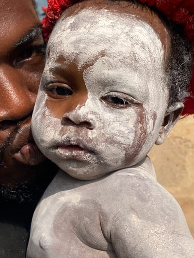 This is a close-up, eye-level shot of a dark-skinned person holding a baby. The baby is partially covered in white powder, with a streak of it running down its face. The baby's left eye is open, looking towards the viewer, while its right eye is partially closed. The person holding the baby has dark skin and a beard, and is leaning in towards the baby. A red object, possibly a headdress or decoration, is visible at the top of the frame. The background is out of focus and appears to be an outdoor environment with a muted brown color. The image was taken in Ufu Esioni, Nigeria.
