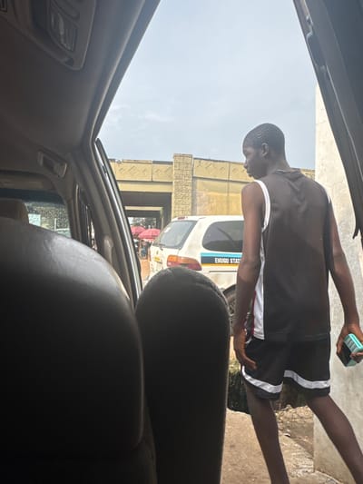A young Black male stands outside a vehicle in Orji, Nigeria. He is wearing a dark sleeveless t-shirt with white trim and black shorts with white trim. He is holding a small, rectangular, light blue object in his right hand. He is looking away from the camera towards a white car with "ENUGU STAT" written on its side. Behind the white car, there are market stalls with red umbrellas and a bridge overhead. The scene appears to be a busy street with traffic and pedestrians. The image is taken from inside another vehicle, looking out through the open door.