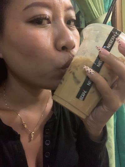 The image is a close-up, medium shot of a person drinking from a to-go cup of iced coffee. The person's face is partially visible, showing their eye, nose, and mouth as they sip from the straw. Their hand, with long, pink nails adorned with black polka dots, is holding the cup, which has a black band with "NESCAFE" printed on it. The person is wearing a black top and a gold necklace with a small pendant. The background is blurred, but appears to be indoors with curtains visible. The image does not provide any information about the location in Jajag, Indonesia.