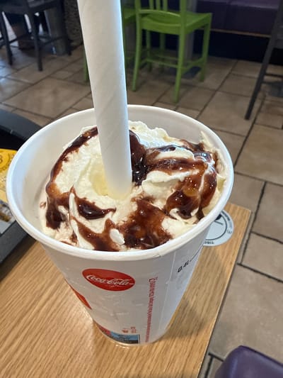 The image shows a close-up of a milkshake in a white disposable cup on a wooden table. The milkshake is topped with whipped cream and drizzled with chocolate syrup. A white paper straw is inserted into the milkshake. The cup has a red Coca-Cola logo. In the background, there are green chairs and tables, suggesting the location is a restaurant or cafe. The floor appears to be tiled. The image quality is good, with clear details of the milkshake and its surroundings. The scene is well-lit, with natural light coming from the side. No people are visible in the image. The overall impression is one of a refreshing treat served in a casual setting. The specific location in Wolfsburg, Germany is not discernible from the image.