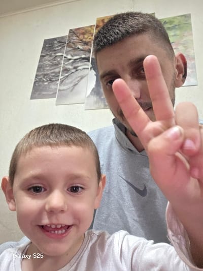 This image depicts a close-up indoor portrait of two individuals, identified as being in the city of Karnalovo, Bulgaria. In the foreground, a young male child, approximately 4-7 years old, is positioned on the left, facing forward with a wide smile revealing his front teeth, including a slight gap between the upper central incisors. He has short, light brown hair and is wearing a light-colored, possibly white or light grey, crew-neck t-shirt. Behind and to the right of the child, an adult male with short, buzz-cut dark hair and visible facial stubble is partially obscured. He is wearing a grey hooded sweatshirt featuring a dark Nike swoosh logo on the left chest. The adult male's right hand is raised in front of his face, making a "peace sign" (V-sign) with his index and middle fingers extended. Both individuals appear to be looking towards the camera. The background consists of a plain, light-colored wall (off-white or light beige). Mounted on this wall are four rectangular canvas prints arranged horizontally, depicting a continuous natural landscape scene. The left two panels feature bare, snow-dusted tree branches, indicative of winter, while the right two panels show trees with autumn foliage, suggesting different seasons or a composite scene. The text "Galaxy S25" is visible in the bottom left corner of the image.