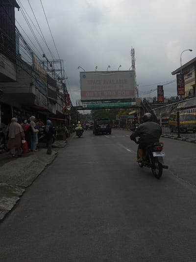 A street scene under a cloudy sky shows various forms of transportation. Pedestrians are gathered on the left sidewalk. A motorbike is driving on the right side of the road. Cars are in the middle lane. Above the street is a large billboard advertising "Space Available" with phone numbers. A pedestrian bridge spans the road with an advertisement. Power lines crisscross above the buildings. A cell tower is visible above the billboard. The photo captures a typical urban street scene.