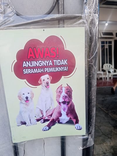 The image shows a sign with three dogs on it, placed on a metal pole. The sign is in Indonesian and warns of aggressive dogs. The dogs depicted are a golden retriever, a labrador, and a pit bull. The sign is partially covered by plastic. The background suggests an outdoor setting, possibly a residential area or a property entrance, with a building and furniture visible in the distance. The sign is attached to a black, textured metal pole. There is no specific indication of the city of Surabaya, East Java, in the image itself.