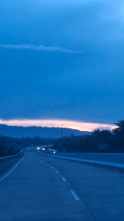 This is a vertical, full-shot photograph of a road at dusk, presumably in or near Seulimeum, Indonesia. The sky is a deep, dark blue, transitioning to a lighter, pinkish-orange hue near the horizon, suggesting a sunset or sunrise. A thin, elongated cloud stretches horizontally across the upper portion of the sky. In the foreground, a paved road curves gently into the distance. White dashed lines mark the lanes of the road. On the left side of the road, a concrete barrier is visible. In the mid-ground, several cars are driving on the road, their headlights illuminated, indicating low visibility. The background is dominated by dark, silhouetted hills and trees. The overall atmosphere is serene and somewhat melancholic due to the fading light and the empty road.