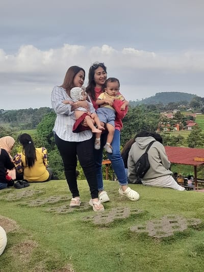Three women and two young children are standing on a grassy hill, likely a tourist site. The woman on the left is wearing a light blue and white striped shirt, black pants, and sandals. She is holding a baby girl with a white top and floral skirt. The woman in the center is wearing a red shirt and black pants and is holding a young boy in a white top and blue shorts. The woman on the right is wearing a gray shirt and jeans and is holding a black bag with a strap. The young boy in the center is holding a toy, possibly a stuffed animal. There are other people sitting on the ground in the background. The environment is rural and green, with hills and trees in the distance. The location is a hilltop in Kecamatan Trawas, Indonesia, with coordinates -7.656 latitude and 112.594 longitude.