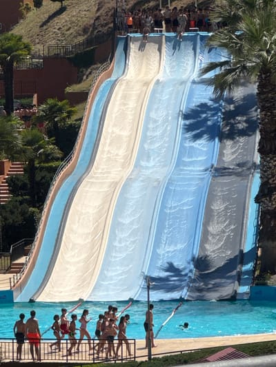 The image depicts a large, multi-lane water slide at a water park. The slide is colored in sections of light blue, white, and grey, with water cascading down its surface. A group of people, predominantly children and teenagers, are gathered at the bottom of the slide, some wading in the turquoise pool. Others are in various stages of ascending the slide or waiting at the top, visible in the background. Palm trees are scattered around the area, and a grassy slope with some trees and stairs is visible behind the slide. The scene suggests a sunny day at a recreational facility. The location is identified as Amarante, Portugal.