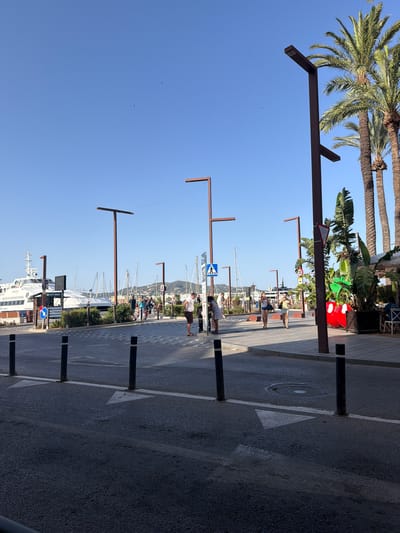 The image depicts a street scene in Ibiza, Spain, likely near the port, as indicated by a large ferry labeled "IBIZA FORMENTERA" docked on the left. The environment is sunny with a clear blue sky. Palm trees are visible, suggesting a warm climate. A paved road with white lane markings and black bollards occupies the foreground. Sidewalks with pedestrians are present in the midground. Several individuals are walking and interacting. On the right side, there is a planter with large tropical plants and a red decorative item resembling Mickey Mouse ears. In the background, sailboats and buildings are visible, with a hilly landscape in the distance. Streetlights with modern designs are strategically placed throughout the scene. The overall atmosphere is one of a lively tourist area.
