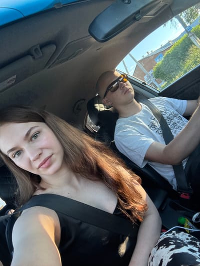 The image shows two people in the interior of a car. In the foreground, a young woman with long auburn hair and green eyes is looking at the camera. She is wearing a black sleeveless top and a seatbelt. To her right, a young man with sunglasses and a white t-shirt is driving. He is also wearing a seatbelt. The car's interior is visible, including the dashboard and a sun visor. Through the car windows, buildings and trees are visible, suggesting an urban or suburban setting. The lighting indicates it is daytime. The location is identified as Кварса, Russia.