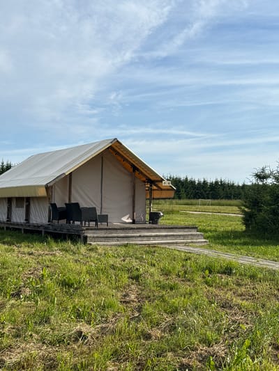 The image shows a glamping tent set up on a wooden platform in a grassy field. The tent has a light-colored fabric covering and is open at the front, revealing a seating area with dark chairs. The platform is made of wooden planks. Beyond the tent, there is a line of trees, and further in the distance, a white structure that resembles a soccer goal can be seen in a field. The sky is blue with wispy white clouds. The sun appears to be high in the sky, suggesting it is daytime, likely late morning or early afternoon. The overall scene evokes a peaceful outdoor setting, possibly a campsite or a rural recreational area. There is a sign with numbers and symbols visible on the tent structure, but the text is too small to read. The image is oriented vertically, with the sky at the bottom and the ground at the top. The location is identified as Zamosh'e (Zaklinskoe S/P), Russia.