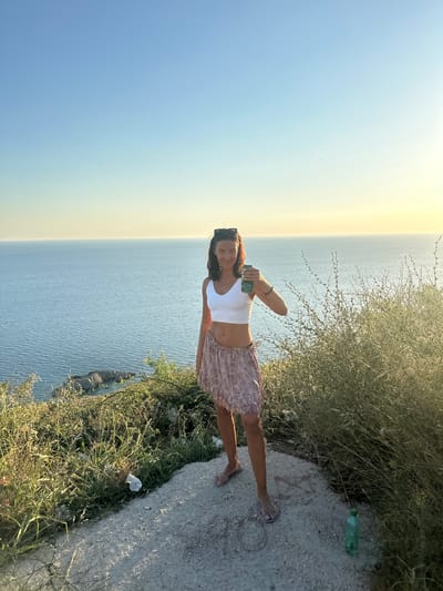 A young woman stands on a rocky cliff overlooking the Adriatic Sea at sunset. She is wearing a white crop top and a patterned skirt. She holds a green can in her right hand. The sky is a gradient of blue and yellow, and the sea is calm. There is sparse vegetation on the cliff, and a green plastic bottle lies on the ground near her feet. The scene is peaceful and serene, with a sense of solitude and tranquility. The location is identified as Donji Katun, Montenegro.