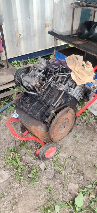 This media file shows a disassembled engine on a red metal stand. The engine is black and appears to be made of metal. It is dirty and covered in oil. There are several hoses and wires attached to the engine. A tan cloth is draped over part of the engine. A rusty flywheel is attached to the engine, and a small red and black wheel is positioned next to the engine on the stand. The scene is outdoors, with dirt, rocks, and green vegetation visible in the background. The lighting suggests it is daytime. The overall impression is that of a discarded or disassembled engine in an outdoor storage area or workshop. No people are visible. The location is noted as Rizunenkove, Ukraine. There is some faint text visible on a white package partially obscured by the engine, which appears to be in Cyrillic script.