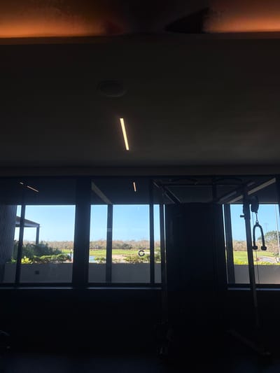 The scene depicts the interior of a facility, likely a gymnasium or fitness center, located in Komchén, Mexico, with a panoramic view through large windows to an outdoor environment.
The interior is dimly lit. The ceiling is dark grey, featuring two illuminated linear light fixtures and a recessed circular fixture, possibly a speaker or another light. A faint, reddish-orange glow is present along the top edge of the ceiling, possibly a reflection or architectural accent lighting. No individuals are present. To the right, a large, dark-colored multi-station cable exercise machine is visible in silhouette, partially obscuring the view through the windows. The brand "Life Fitness" is discernible on its side. Cable attachments, including a rope handle, hang from the machine. The floor and lower walls are dark and largely featureless due to shadowing.
Through the multi-pane windows, a bright, clear blue sky is visible. The exterior landscape features a body of water in the middle ground, reflecting the sky. Beyond the water, an extensive, manicured grassy area, characteristic of a golf course, stretches into the distance, interspersed with trees and shrubs under a clear sky. In the immediate foreground outside the windows, a landscaped area with green foliage and a low concrete wall is visible. To the far left, an architectural structure with vertical slatted panels is partially seen. A white circular object with a vertical line, possibly a sign or marker, is visible on the grassy terrain in the mid-ground. No human activity or movement is observed in the exterior scene.
