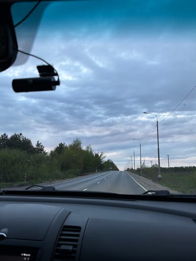 View from inside a car driving on a two-lane road in Слобода, Russia. The road is flanked by trees and utility poles. The sky is overcast. A dashcam is mounted on the windshield. The car's dashboard and air vents are visible. No people are visible.