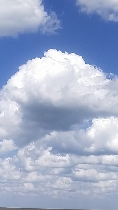 This is a close-up, vertical photograph of a bright blue sky filled with a variety of white and gray clouds. The clouds range in texture from fluffy and wispy to dense and shadowed. The bottom of the frame shows a sliver of a muted, light brown horizon, possibly land or water. There are no people or man-made objects visible in the image. The photograph appears to have been taken on a sunny day. The location is specified as Озёрный ауылдық округі, Kazakhstan.