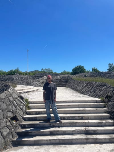 The image depicts a man standing on a wide, stepped staircase in Barutana, Montenegro. He has pink hair, wears glasses, a grey t-shirt, light blue jeans, and brown shoes. The staircase is flanked by stone walls on both sides, ascending a grassy slope lined with trees. A light pole extends into a clear, blue sky. The scene is brightly lit. There's a small niche in the stone wall near the bottom of the stairs. The location is characterized by stone and natural elements.