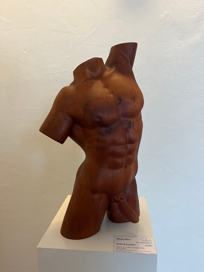 The image displays an indoor scene within the Bellarte gallery, located in Staudernheim, Germany. A dark reddish-brown wooden sculpture of a male torso is centrally positioned on a white rectangular pedestal. The sculpture, rendered in mahogany wood, depicts a muscular physique, complete with defined pectoral muscles, a six-pack abdomen, and explicitly detailed male genitalia. It is headless and armless, with smooth, unfinished surfaces at the neck and shoulder/upper arm junctions.

To the lower right of the sculpture, a white informational label is affixed to the pedestal. The label identifies the artist as "Wayan Wina," the artwork title as "Kraftvoll & Schön" (Powerful & Beautiful), and the material as "Mahagoniholz" (Mahogany wood) with a height of "40 cm." A price of "€ 490,-" is also displayed, along with the "bellarte" logo and website "www.online-bellarte.de." The background consists of a plain, off-white textured wall. The scene is evenly lit.
