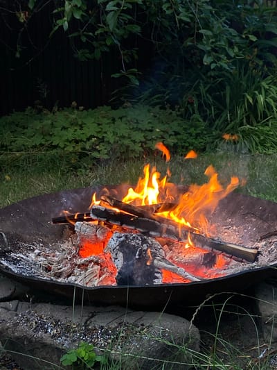 An outdoor fire pit contains burning logs and embers, illuminating the surrounding area with a warm glow. The fire pit is a dark, metal bowl set on the ground. The ground around the fire pit is covered with grass, stones, and scattered ash. In the background, lush green foliage creates a natural backdrop, suggesting a rural or garden setting. The scene is dimly lit, with the fire being the primary light source. There are no people or animals visible in the image. The location in Лышчыцкі сельскі Савет, Belarus, implies a potentially rural or suburban environment common in such Belarusian regions.
