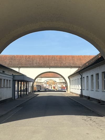 The image displays an architectural scene in Bad Nauheim, Germany, framed by a large, light-colored, textured archway in the immediate foreground. A paved road or path occupies the lower half, extending towards the background. On the left, a white-walled building features a modern-style entrance with dark-framed glass panels and a dark canopy. On the right, a parallel white-walled building presents a series of rectangular windows. Both flanking buildings possess reddish-brown tiled roofs. Across the midground, a substantial white-walled structure, also topped with a reddish-brown tiled roof, bridges the gap between the side buildings. This structure contains a smaller, central archway. Through this archway, an ornate architectural facade with a prominent central decorative element is visible in the background, flanked by walls adorned with multiple colorful murals or large artworks. Indistinct objects are also present in this distant area. The overhead sky is clear and blue. No individuals, specific actions, or interactions are depicted within the frame.