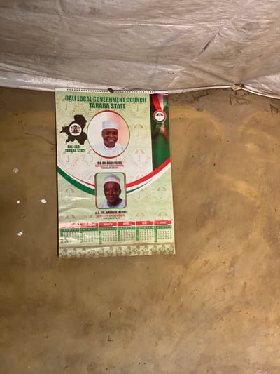 A calendar is hanging on a rough, textured wall. The calendar features portraits of two men and a map of Nigeria with a small emblem on it. Text on the calendar reads "BALI LOCAL GOVERNMENT COUNCIL TARABA STATE," "H.E. DR. AGBU KEFAS EXECUTIVE GOVERNOR, TARABA STATE," and "H.E. DR. AMINU A. ALKALI DEPUTY GOVERNOR, TARABA STATE." Below the portraits are calendar grids for February, March, April, May, and June. The wall appears to be made of mud or a similar material, with visible imperfections and discoloration. Above the calendar, a white, tarp-like material is stretched across the ceiling. The lighting suggests an indoor environment with some natural light.