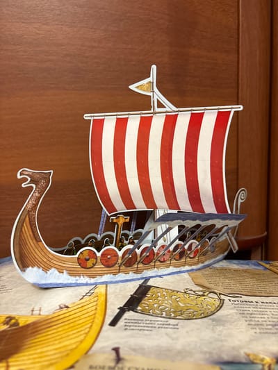 This is a close-up, slightly angled shot of a detailed pop-up book depicting Viking longships. The foreground features the front section of a Viking ship with a large red and white striped sail. Shields are visible along the side of the ship, and small figures of Vikings can be seen standing on deck. The ship is rendered with intricate details, including the wooden texture of the hull, the waves of the sea below, and a decorative dragon head at the prow.
Behind the primary ship, another pop-up element is visible, appearing to be another longship rendered in yellow. The background consists of a flat, dark brown surface, likely the cover or a page of the book itself, with some text in Russian. The text, partially visible and legible, includes phrases like "Готовы к напад" (Ready for attack) and mentions "Викинги" (Vikings), suggesting the book's theme. The lighting is bright, highlighting the vibrant colors and detailed artwork of the pop-up elements. The scene appears to be indoors, with the focus solely on the book's content.