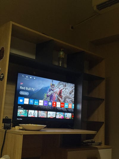 A flat-screen television is the primary subject, displaying the "Red Bull TV" interface with a motocross rider as the background image. Below the Red Bull logo and "Watch Now" button, various streaming service icons are visible, including TV, Netflix, Prime Video, Disney+, YouTube, VOYO, HBO Max, and Arte. Below these, a "Netflix Recommended" row shows several movie posters. The TV is situated on a wooden shelf in what appears to be a room. To the left of the TV, a black power adapter is plugged into a wall outlet. In front of the TV, a white bowl containing food rests on the shelf. To the right, on a lower shelf, two remote controls are visible. The overall environment is dimly lit, with shadows cast by shelving units. The image is taken from a slightly high angle, looking down at the television. The location is described as being in the city of Peshtera, Bulgaria.